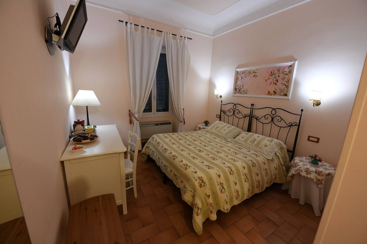Una camera da letto con un letto, una scrivania, una sedia, una finestra con tende, una TV a parete e un comodino con una lampada.