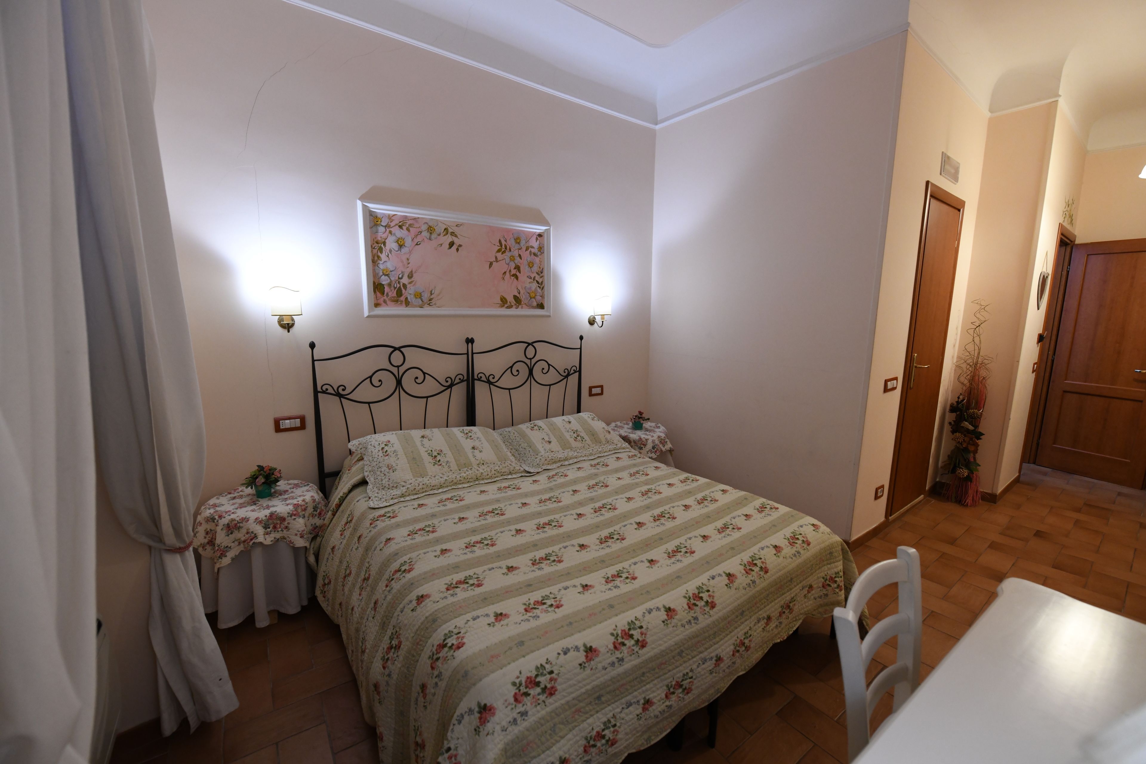 A bedroom with a bed, two bedside tables, a nightstand, a wall-mounted light, a floral painting, and a wooden door.