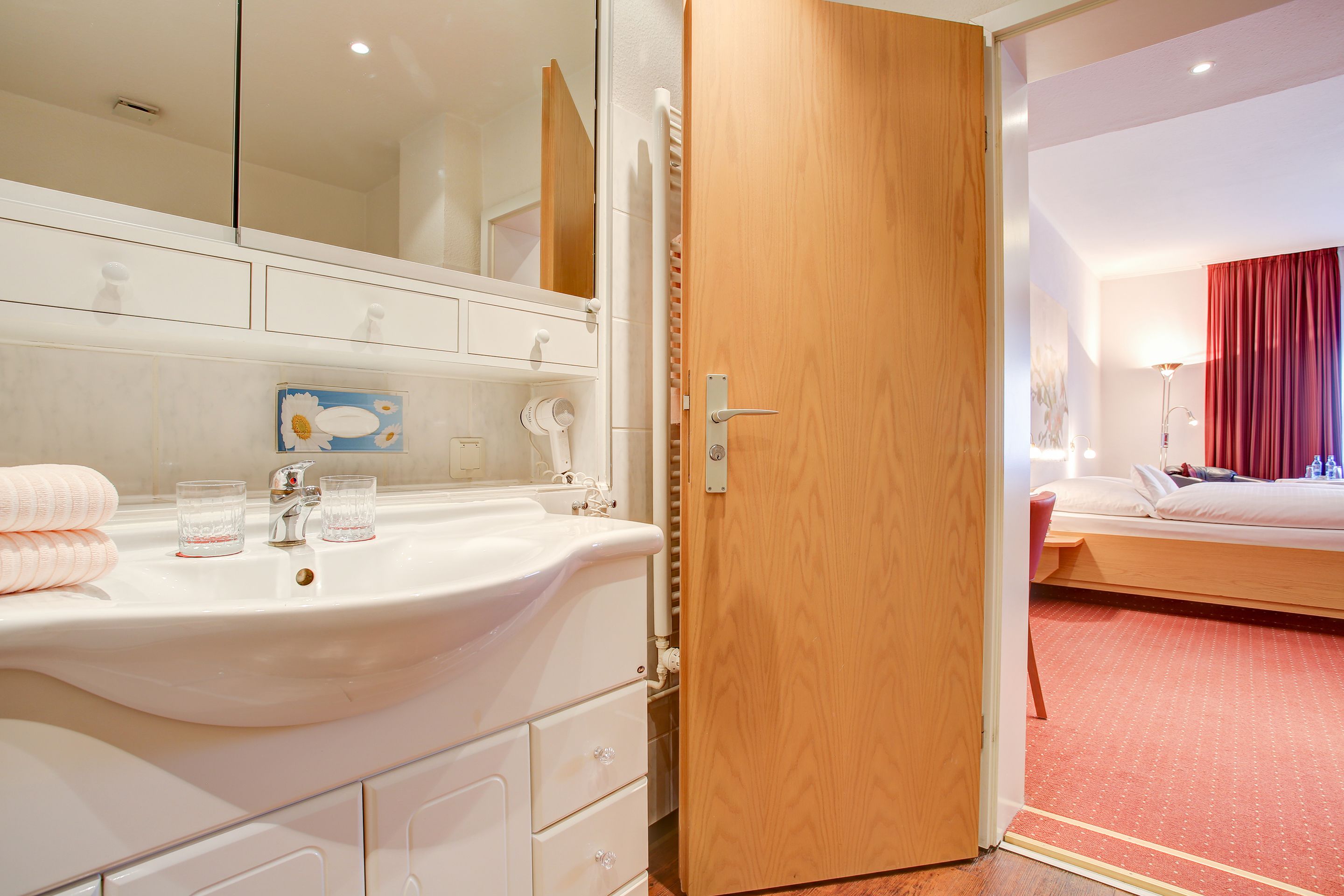 A hotel room with a white sink, a mirror, a wooden door, a bed with a red curtain, and a red carpeted floor.