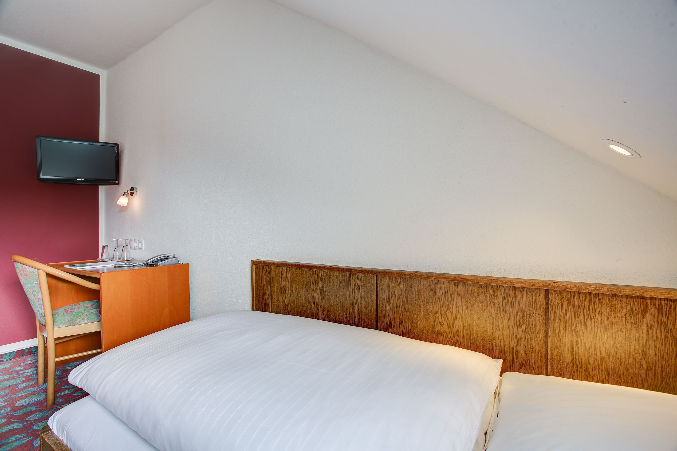 A hotel room with a bed, a desk with a chair, a television mounted on the wall, and a small table with a lamp.
