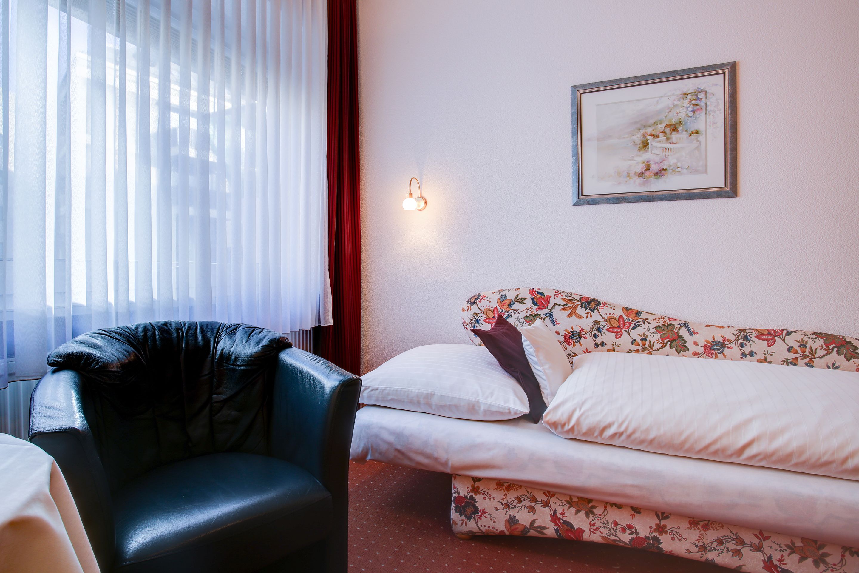 A room with a black leather chair, a floral-patterned sofa, a framed artwork on the wall, and a window with sheer curtains.
