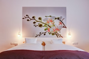 A neatly made bed with white linens and a red throw blanket, a wall-mounted orchid painting, and bedside lamps. - Hotel Consul (Kiel)
