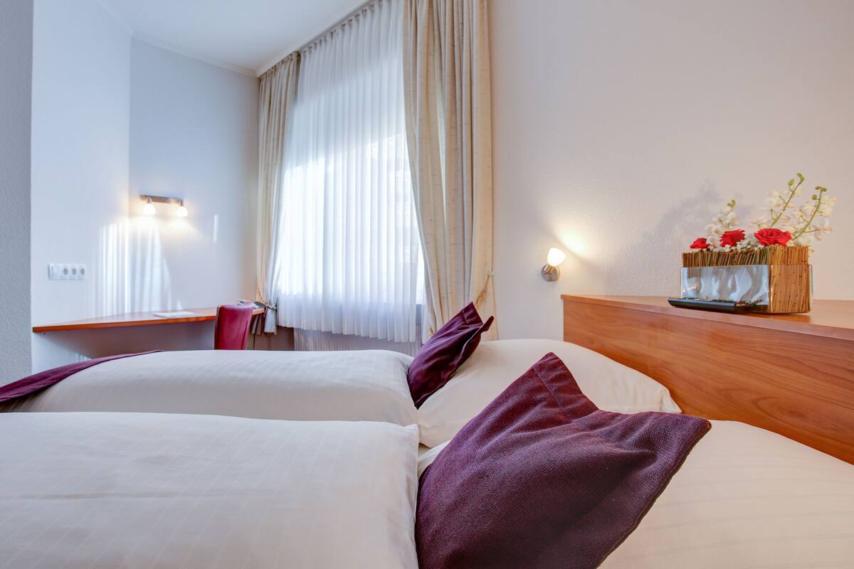 Quarto de hotel com cama, mesa de cabeceira de madeira, escrivaninha e janela com cortinas.