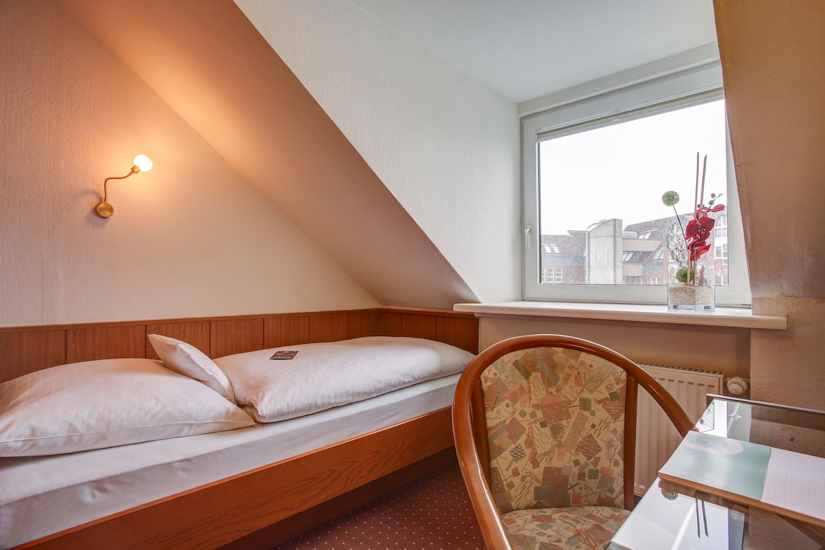 Quarto pequeno e acolhedor com uma cama de solteiro, uma cadeira, uma mesa e janela com vista para edifícios.