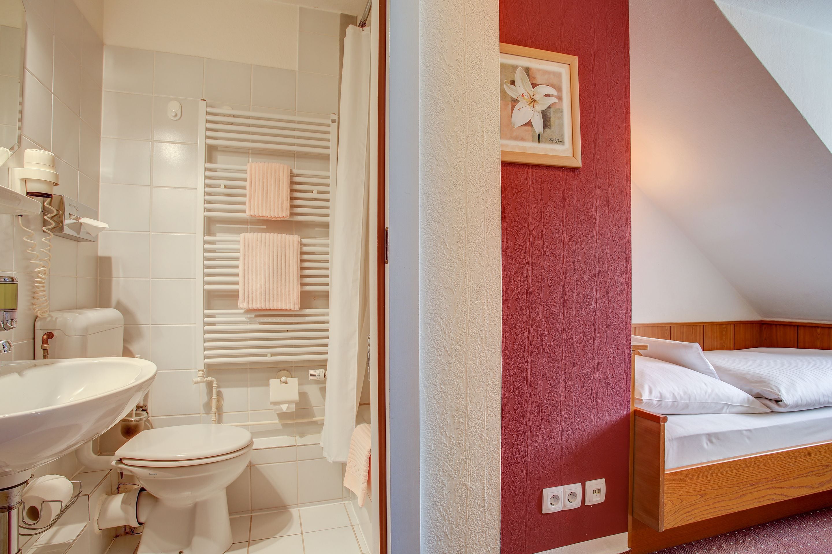 A bathroom with a sink, toilet, and shower. There is a bed with white bedding and a framed picture on the wall.