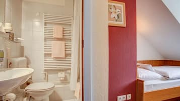 A bathroom with a sink, toilet, and shower. There is a bed with white bedding and a framed picture on the wall.