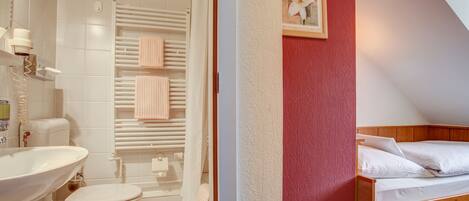 Une salle de bain avec un lavabo, des toilettes et une douche. On y trouve un lit avec du linge de lit blanc et un tableau encadré au mur.