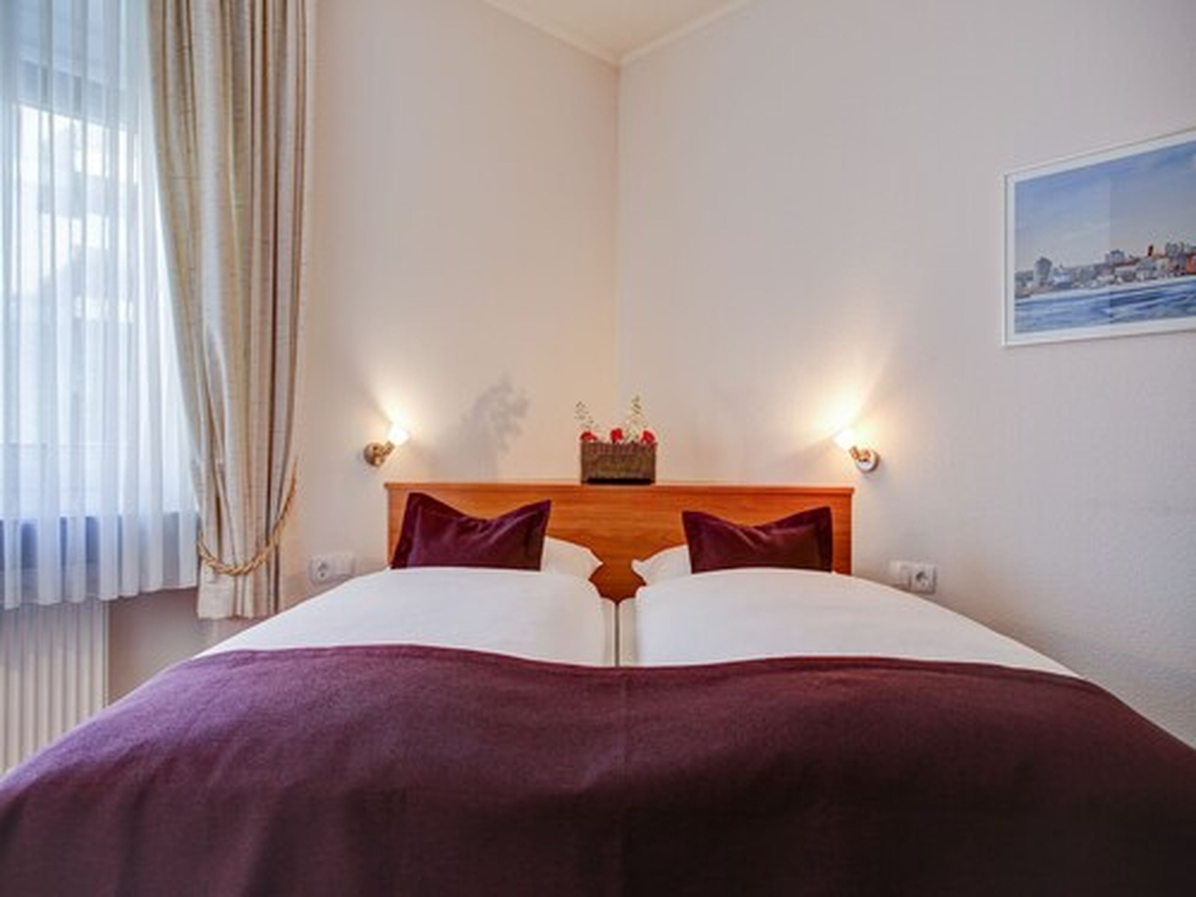 Habitación de hotel con cama, dos lámparas de noche, mesita de noche con un adorno y una ventana con cortinas.