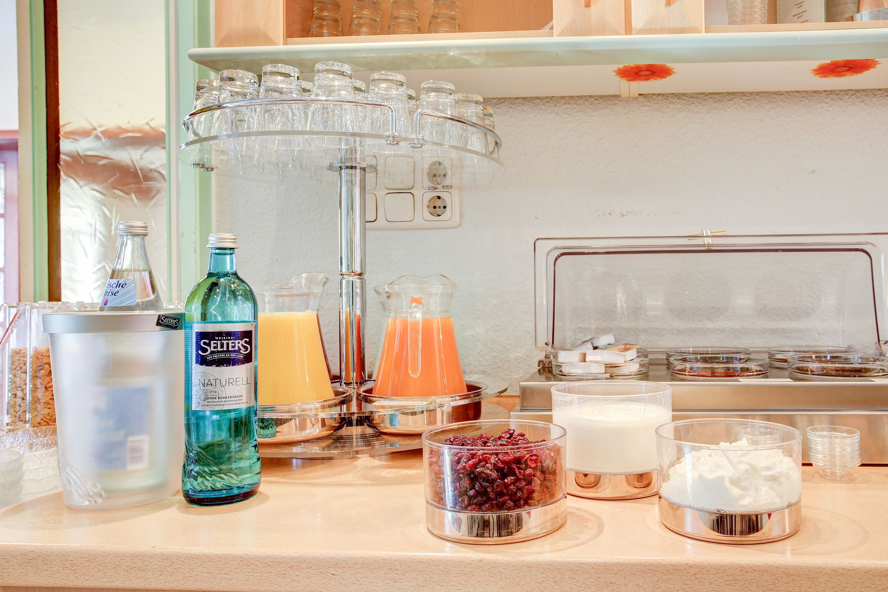 Un buffet de petit-déjeuner proposant diverses boissons et aliments, notamment une bouteille d’eau gazeuse, un pichet en verre, ainsi que des pots de yaourt et des barquettes de fruits rouges.