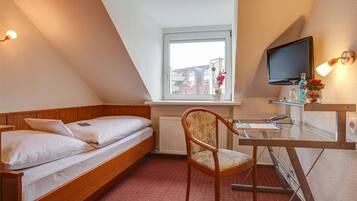 A small, cozy room with a wooden bed, a desk with a chair, a television, and a window with a view of buildings.