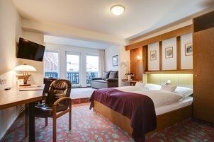 Habitación de hotel con cama, escritorio con silla, televisor montado en la pared y balcón con vistas a edificios.