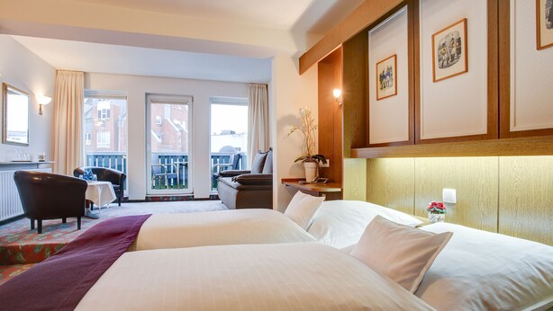 A hotel room with two beds, a desk, a chair, and a balcony with a view of buildings. - Hotel Consul (Kiel)