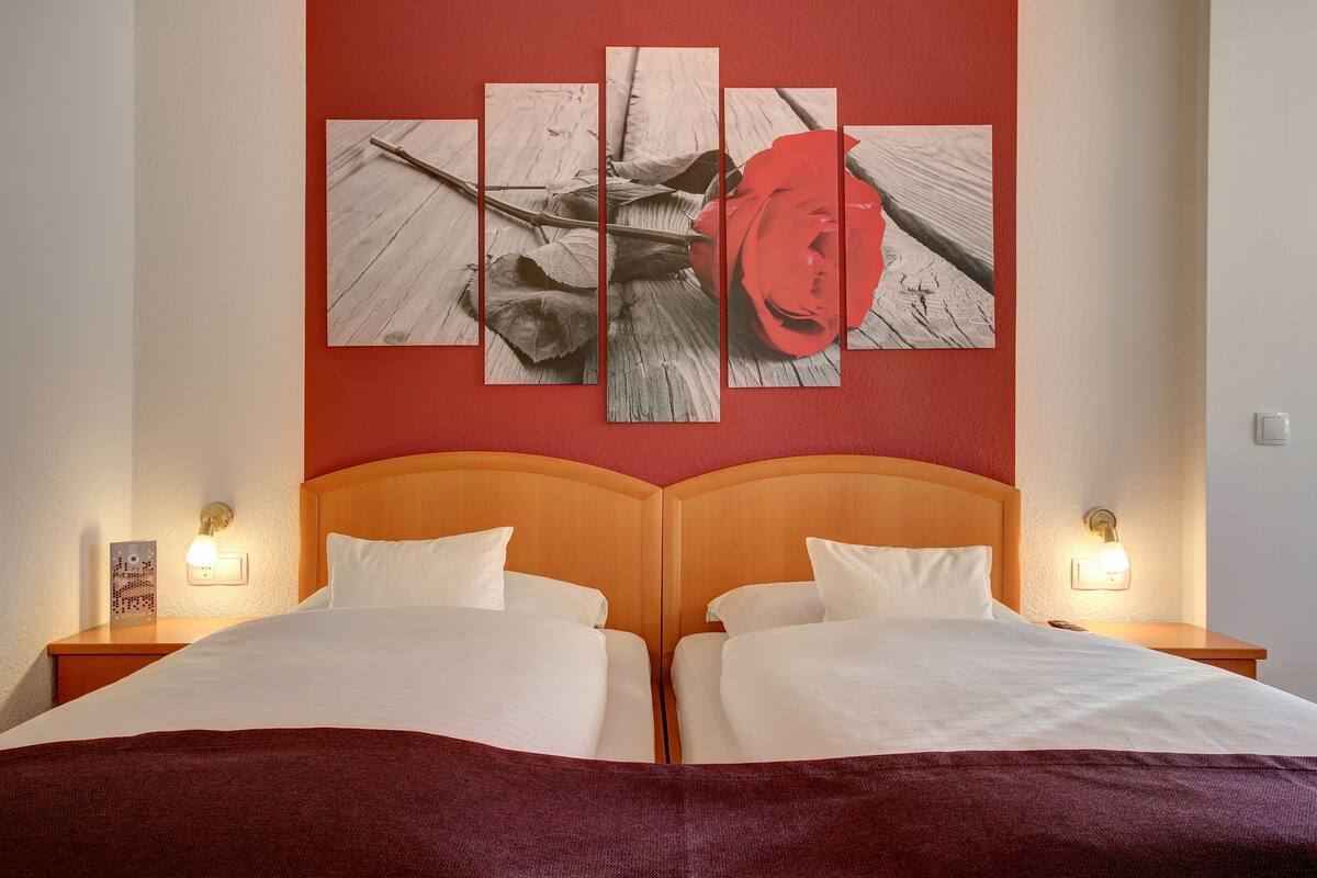 Quarto de hotel com uma cama grande, duas mesas de cabeceira e uma obra de arte na parede retratando uma rosa.
