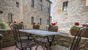 Un bâtiment en pierre avec une table et des chaises dans une cour, le tout entouré de plantes en pot aux fleurs rouges.
