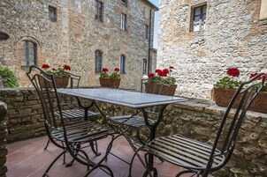 Edificio de piedra con una mesa y sillas en el patio, rodeado de macetas con flores rojas.