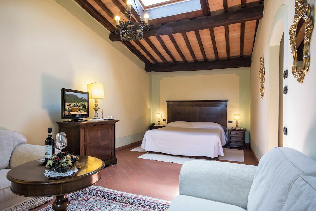 Una habitación de hotel con una cama, un sofá, una mesita con una botella y un vaso, una lámpara y un candelabro.
