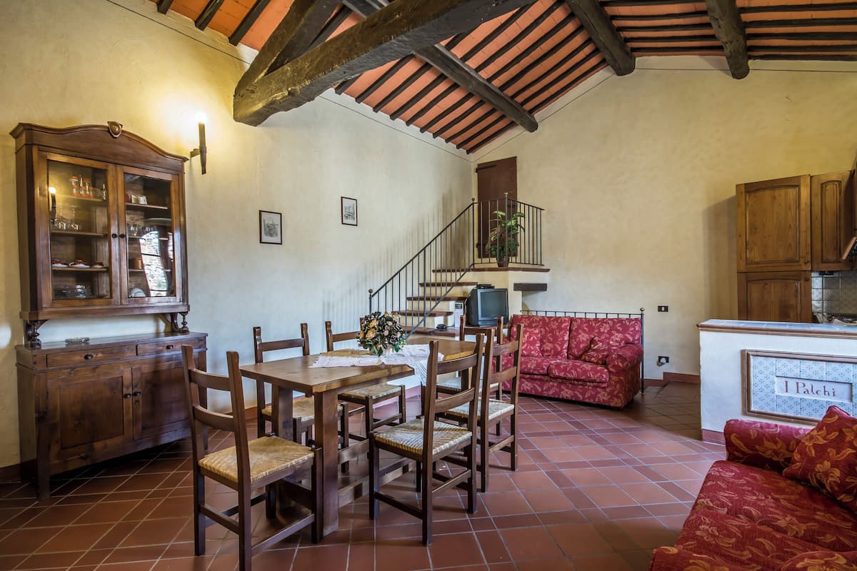 Un comedor con mobiliario de madera, un sofá rojo con estampado y una zona de cocina con armarios de madera.