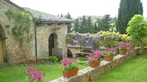 Un bâtiment en pierre doté d’une entrée en arc de cercle, un jardin avec des plantes en pot et une pergola couverte de plantes grimpantes.