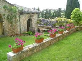 Un edificio de piedra con una entrada en arco, un jardín con flores en macetas y una pérgola con hiedra colgante.