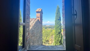 Une porte ouverte donne sur une vue d’un bâtiment historique, avec un toit recouvert de tuiles et une cheminée, le tout entouré de verdure et surplombé par un ciel bleu clair.