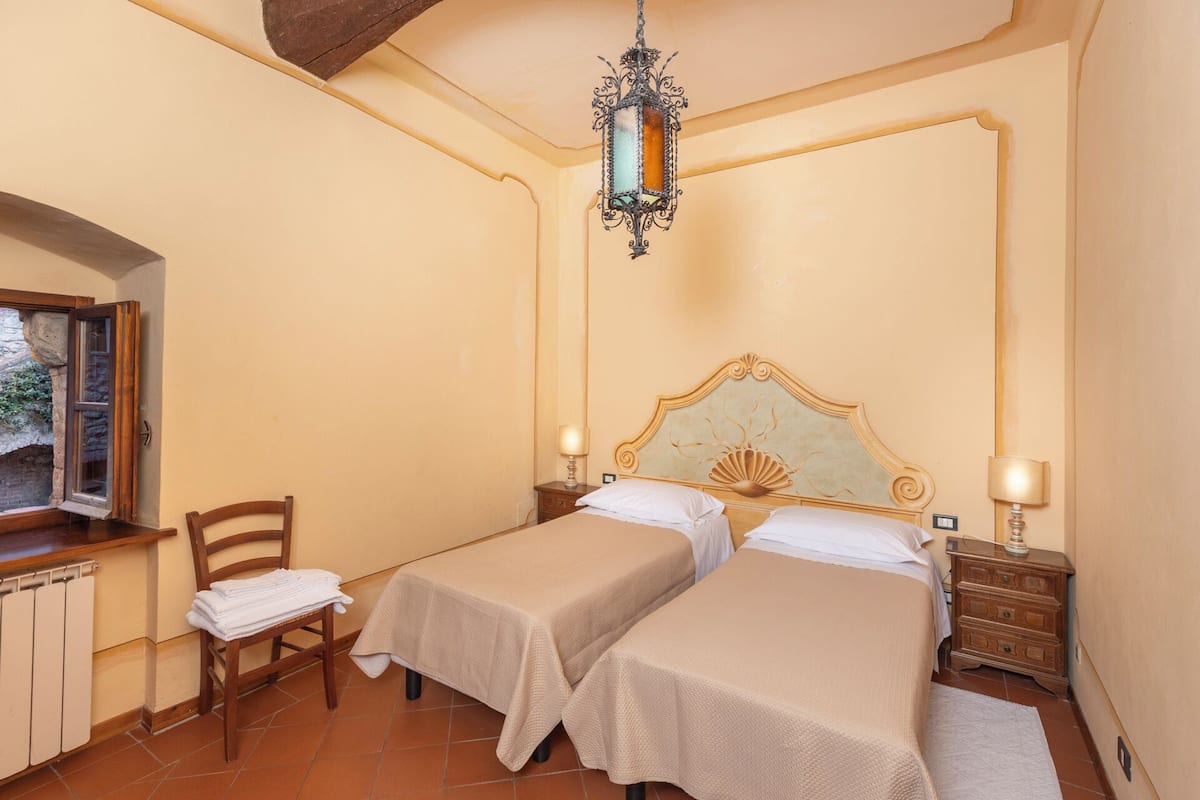 Una habitación con dos camas, una silla de madera, una mesa y una ventana con vista a follaje.