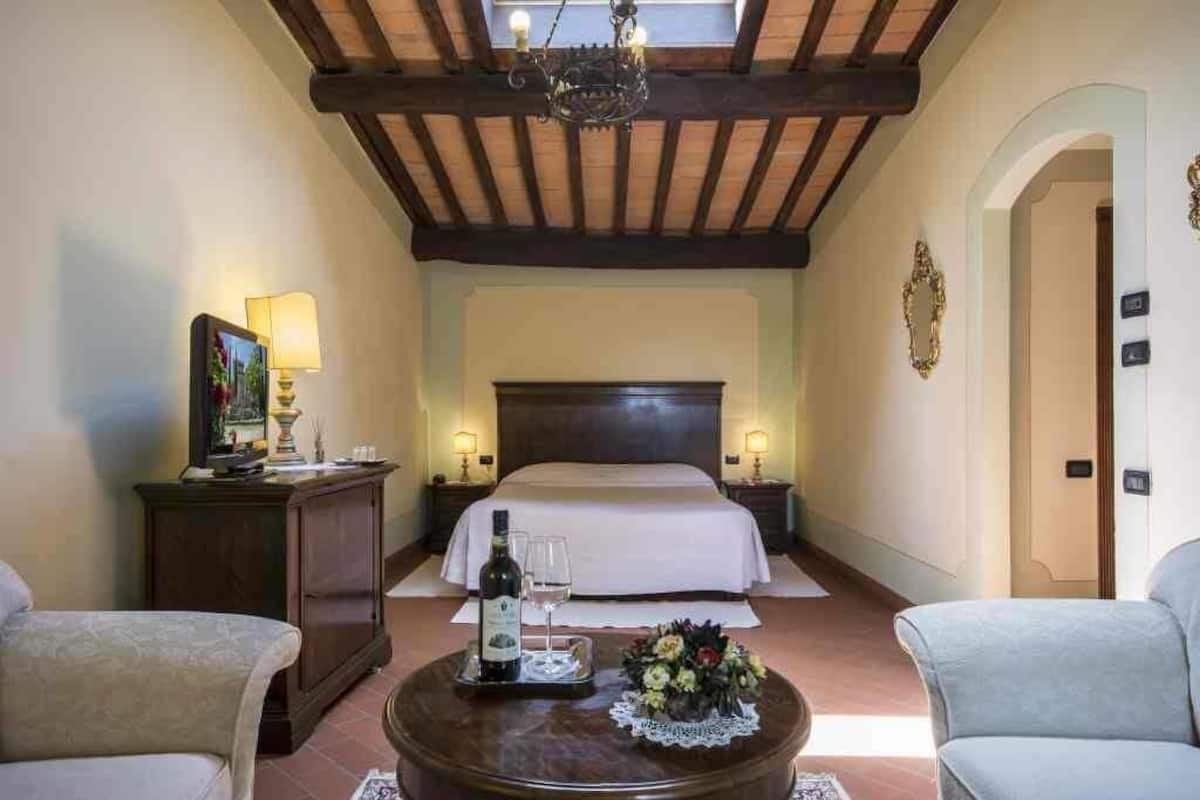 Habitación de hotel con una cama, una mesita de noche, una lámpara, una mesa con una botella y vasos, y una silla.