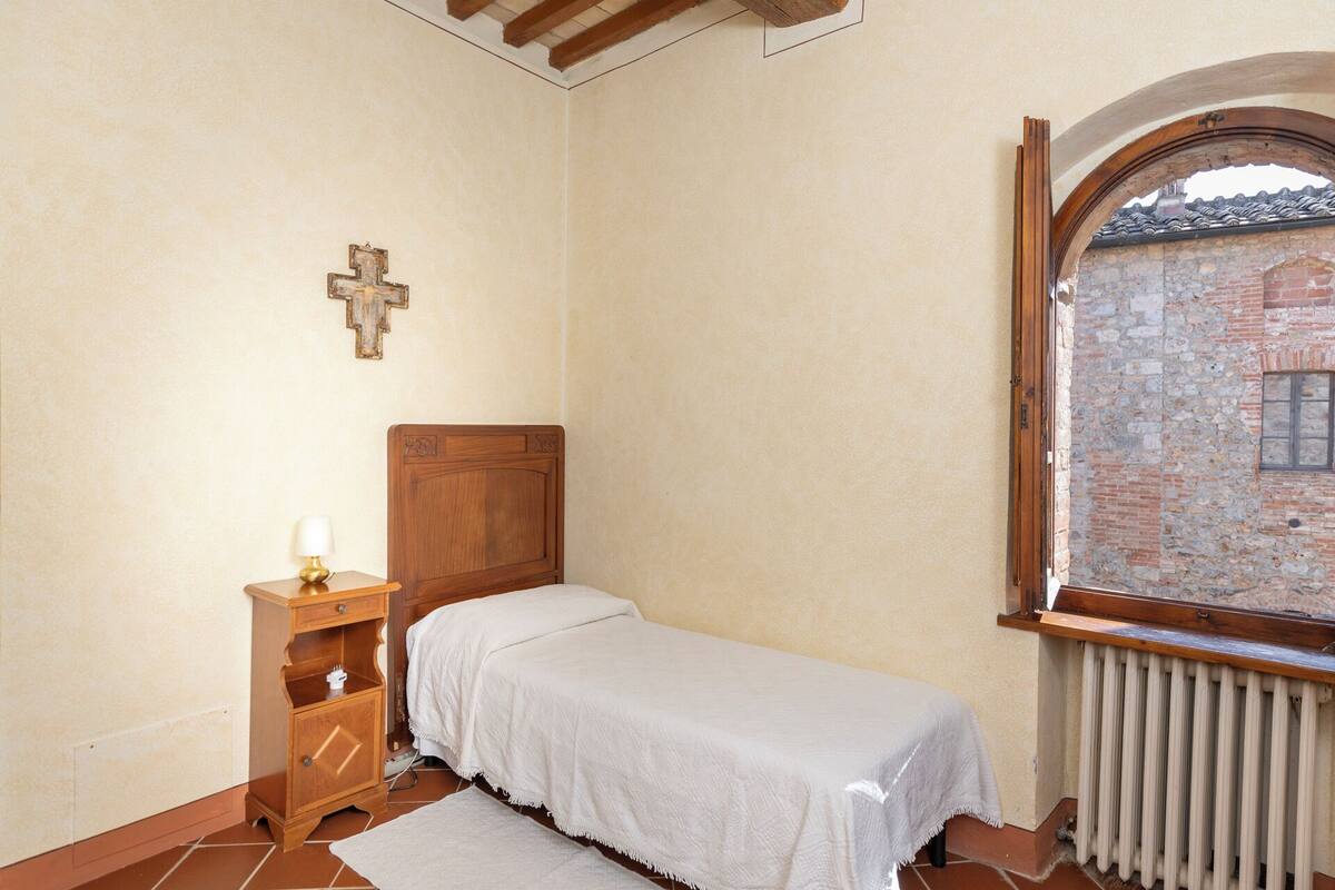 Un dormitorio con una cama de madera, una mesita de noche, una cruz en la pared y una ventana con vistas a un edificio de ladrillo.