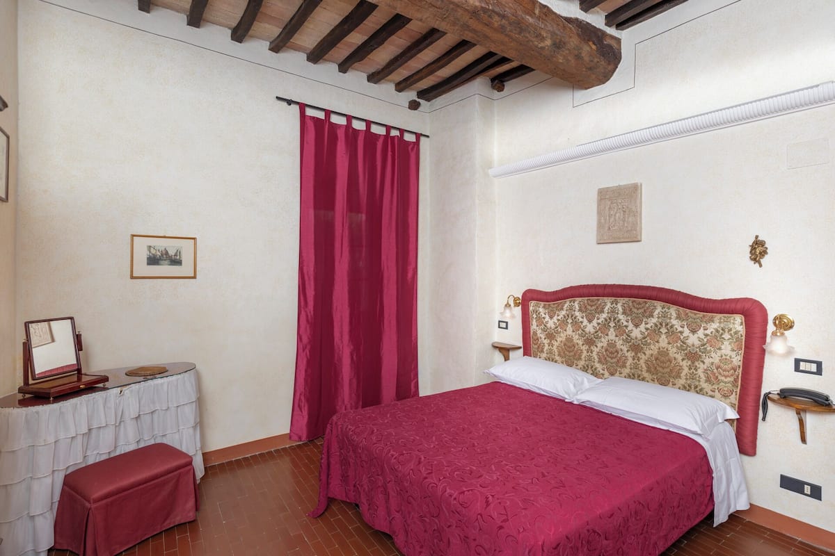 Un dormitorio con techo de madera, una cama con cabecera floral, una cortina roja, una mesita de noche con lámpara y un pequeño marco de fotos en la pared.