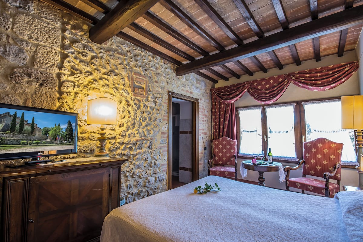 Una habitación con paredes de piedra, una cama, un televisor, una mesa con una botella y flores, y sillas tapizadas en rojo.