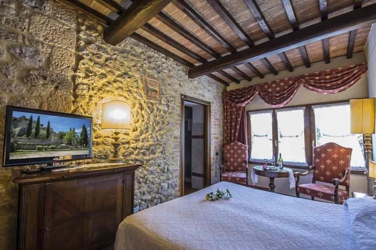 A room with a stone wall, wooden beams, a bed, a television, a chair, a table, and a window with curtains.