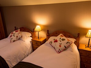A room with two beds, each with a wooden headboard and a nightstand with a lamp. There are floral-patterned pillows on the beds.