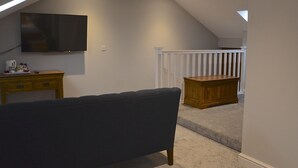 A room with a TV mounted on the wall, a wooden dresser, a dark sofa, and a white railing in the background. - The Barrel Inn (Hope Valley)