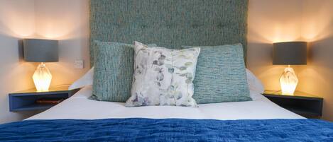 A neatly made bed with a blue blanket, a green upholstered headboard, and two bedside lamps.