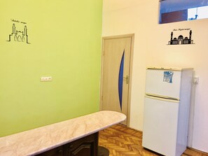 Une petite pièce avec une porte en bois, un réfrigérateur blanc, un comptoir en marbre et des œuvres d’art murales représentant une mosquée.