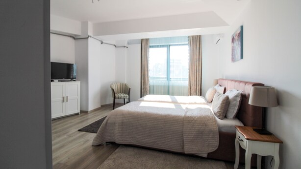 Ein modernes Schlafzimmer mit einem großen Bett, einem Nachttisch, einem Sessel, einem Fernseher und einem Fenster mit Vorhängen.