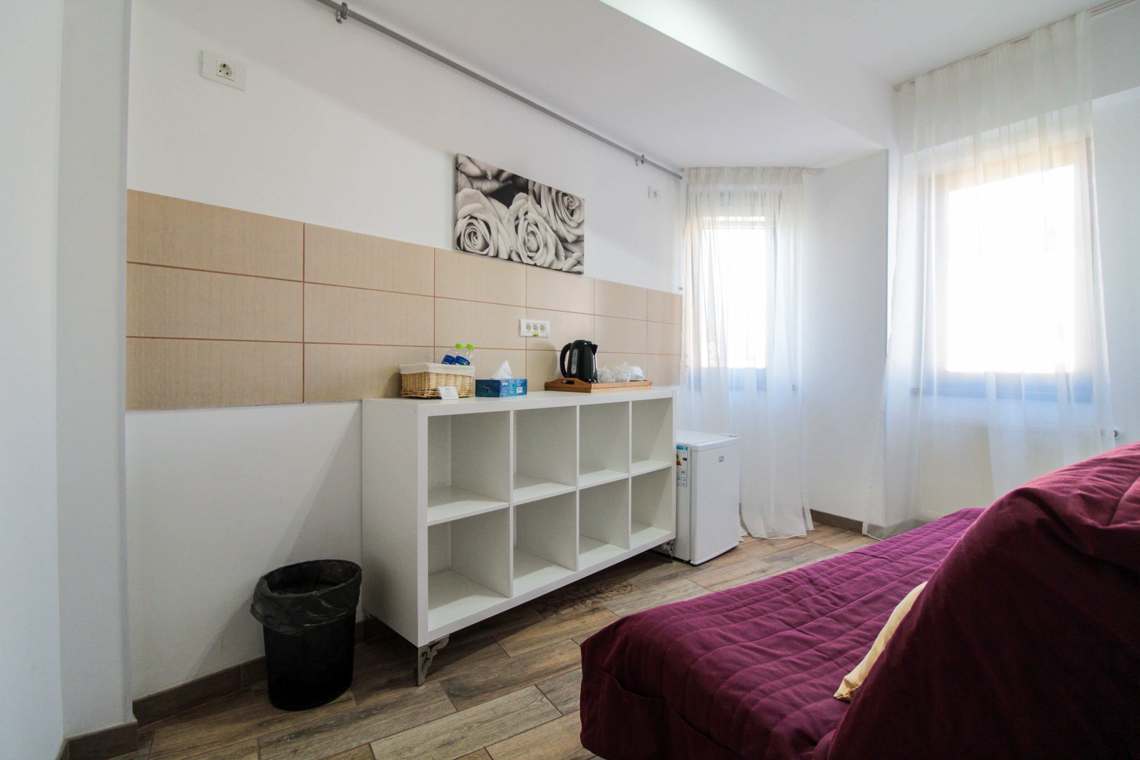 A room with a bed, a white shelf unit, a small refrigerator, a trash bin, and a window with sheer curtains.