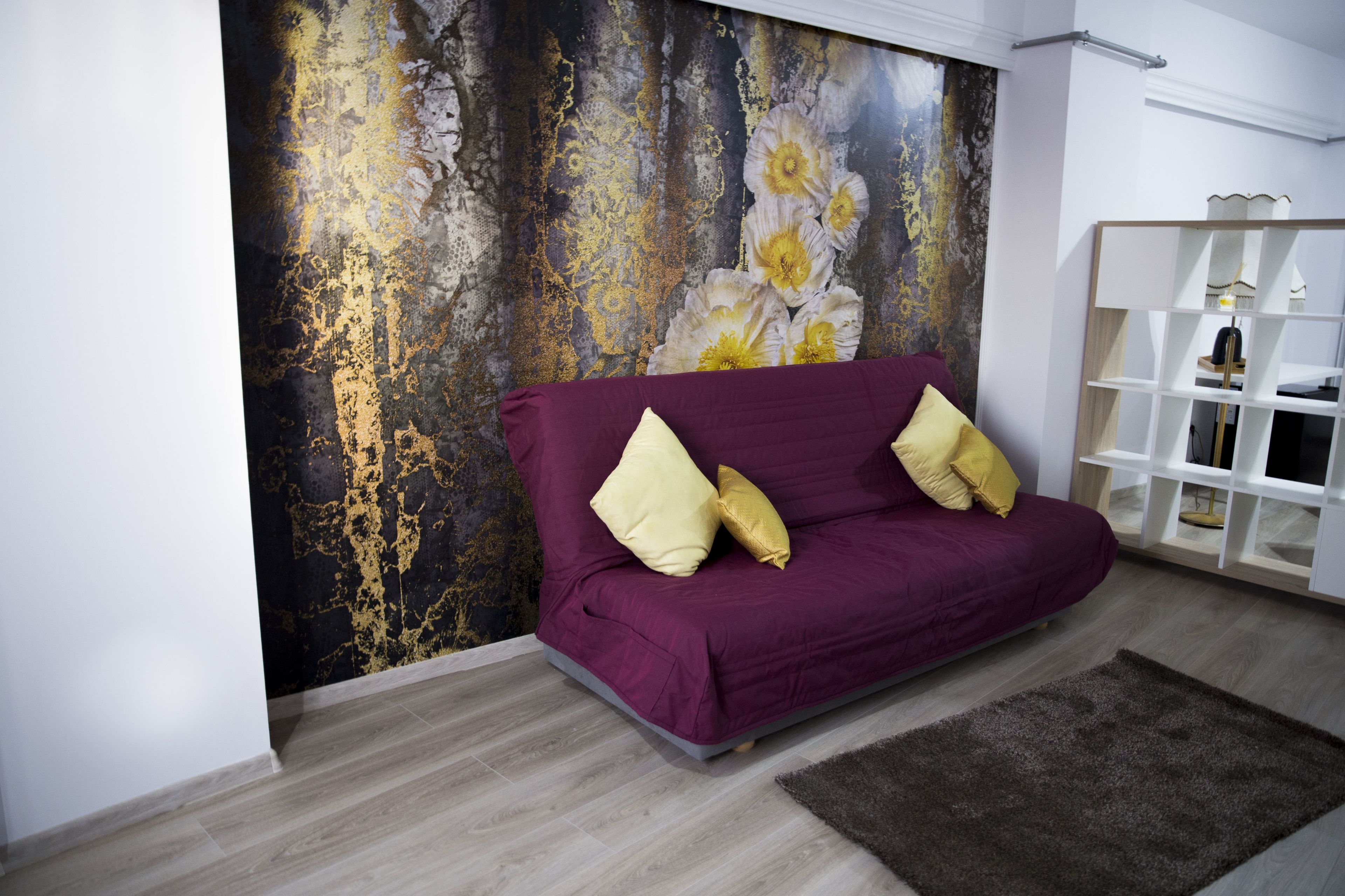 A modern living room with a purple sofa, a decorative wall with a floral pattern, and a white shelving unit.