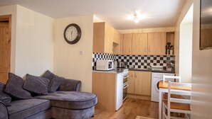 A compact living space with a sofa, a dining table, a kitchen area including a microwave and a washing machine, and a clock on the wall. - 2 Varis Apartments (Forres)