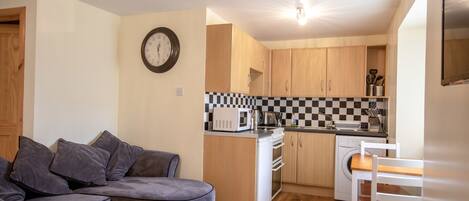A compact living space with a sofa, a dining table, a kitchen area including a microwave and a washing machine, and a clock on the wall.