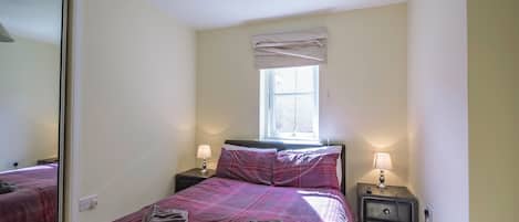 A bedroom with a bed, two bedside tables with lamps, a window with a curtain, and a mirror on the wall.