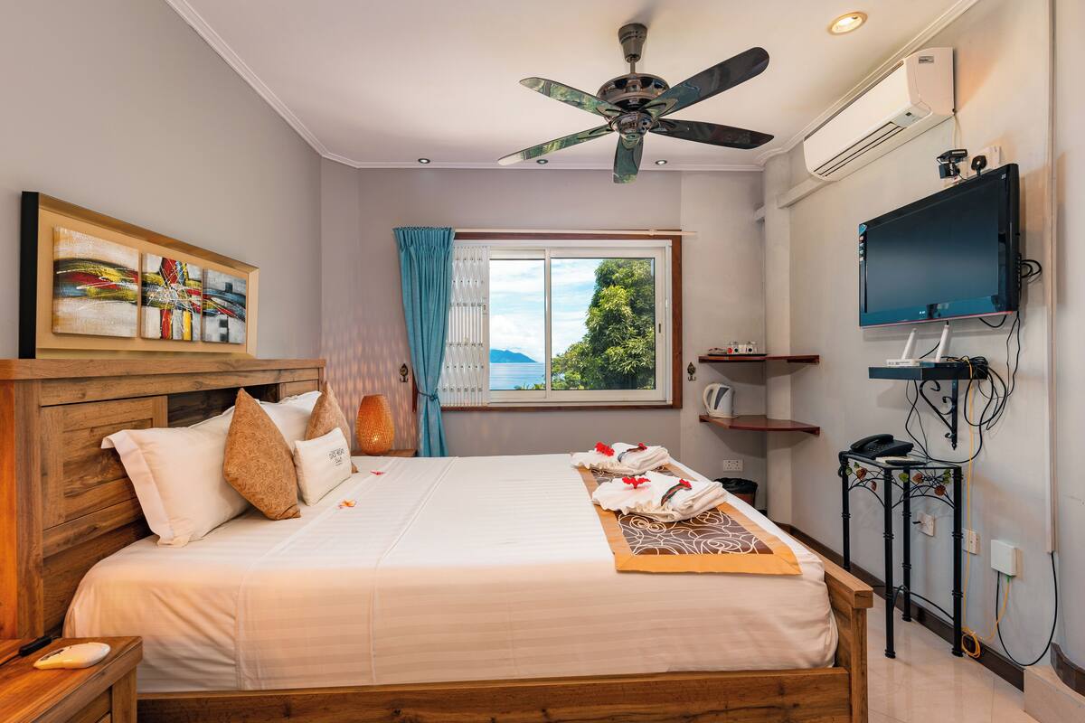 Une chambre d’hôtel avec un lit, un ventilateur de plafond, une télévision, une fenêtre avec des rideaux et un tableau au mur.