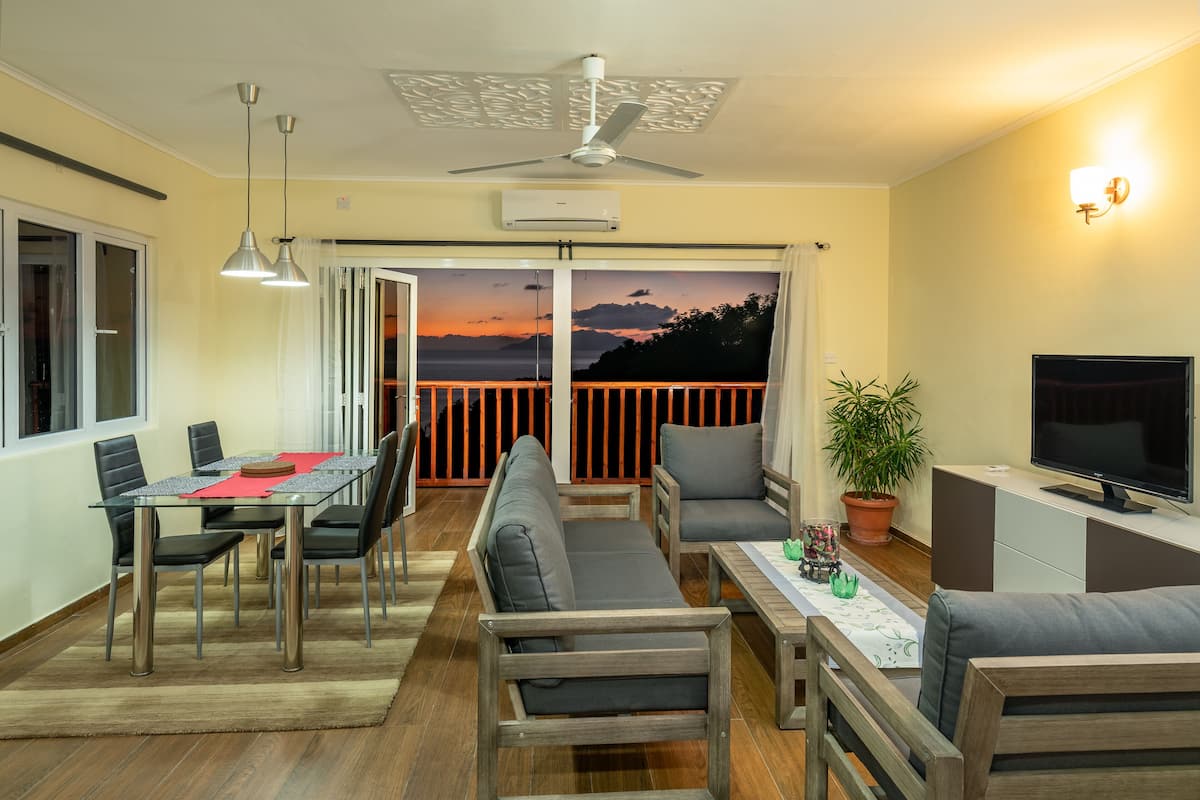 Un salon moderne avec une table à manger en verre, des canapés, un téléviseur et un balcon offrant une vue sur le coucher du soleil.