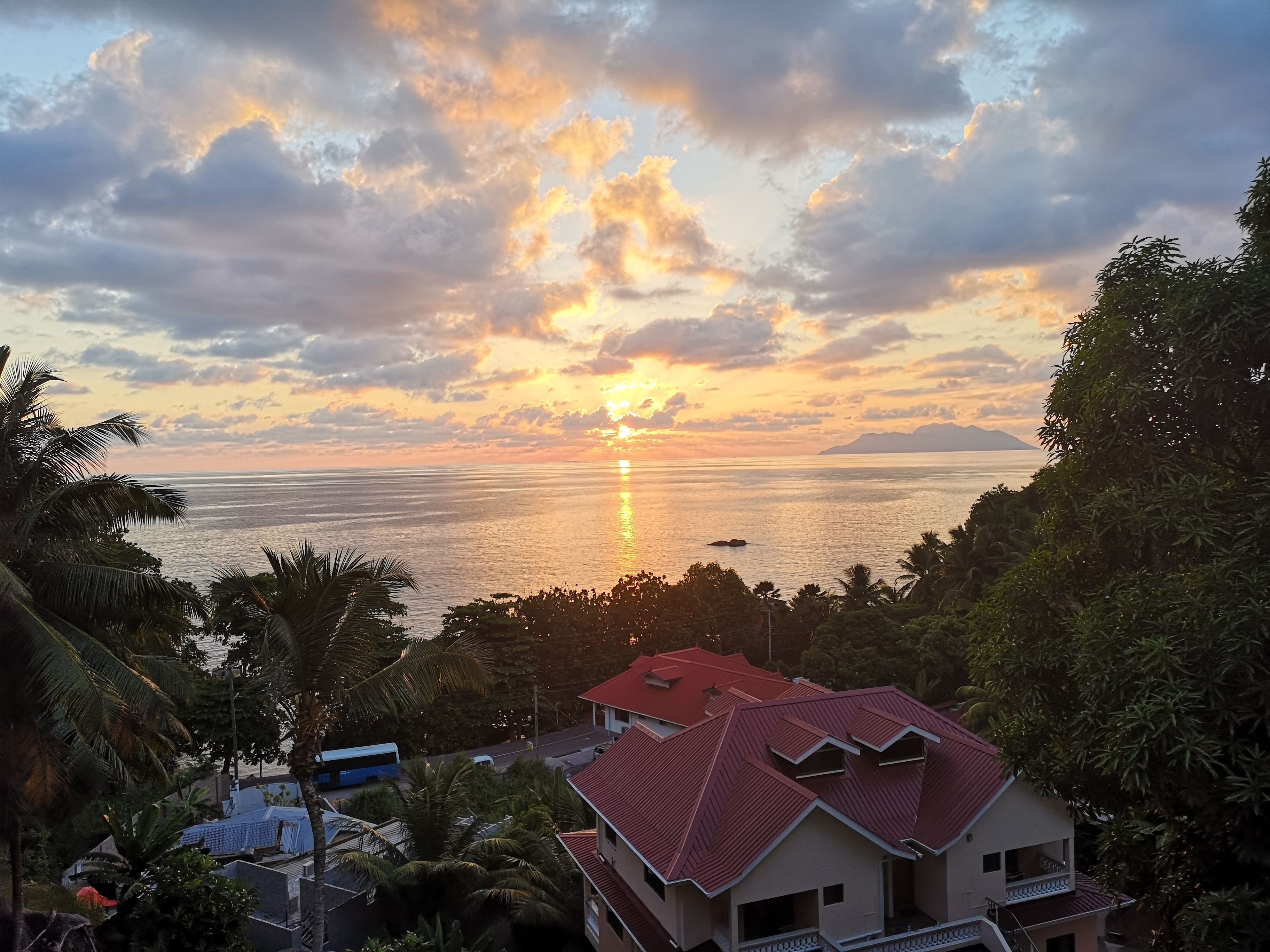 Vista costera con atardecer, una casa de techo rojo y palmeras.