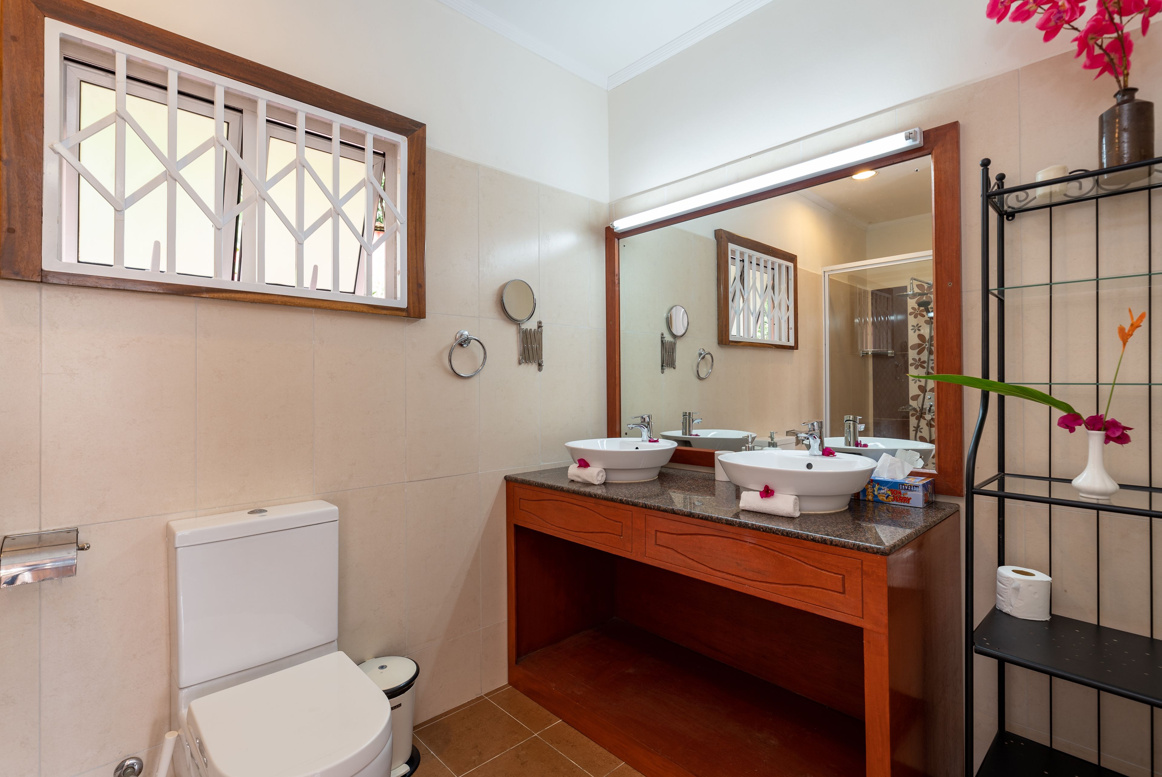 Une salle de bain avec deux lavabos, des toilettes, une fenêtre ornée d’une grille décorative et un miroir au-dessus des lavabos.