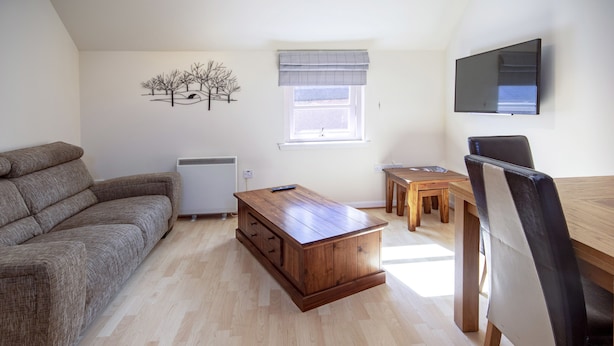 A living room with a sofa, a wooden coffee table, a dining table with chairs, a TV mounted on the wall, and a window with a view.