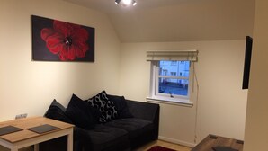 A living room with a black sofa, a wooden table, a red rug, a window with a view of buildings, and a large red flower painting on the wall.