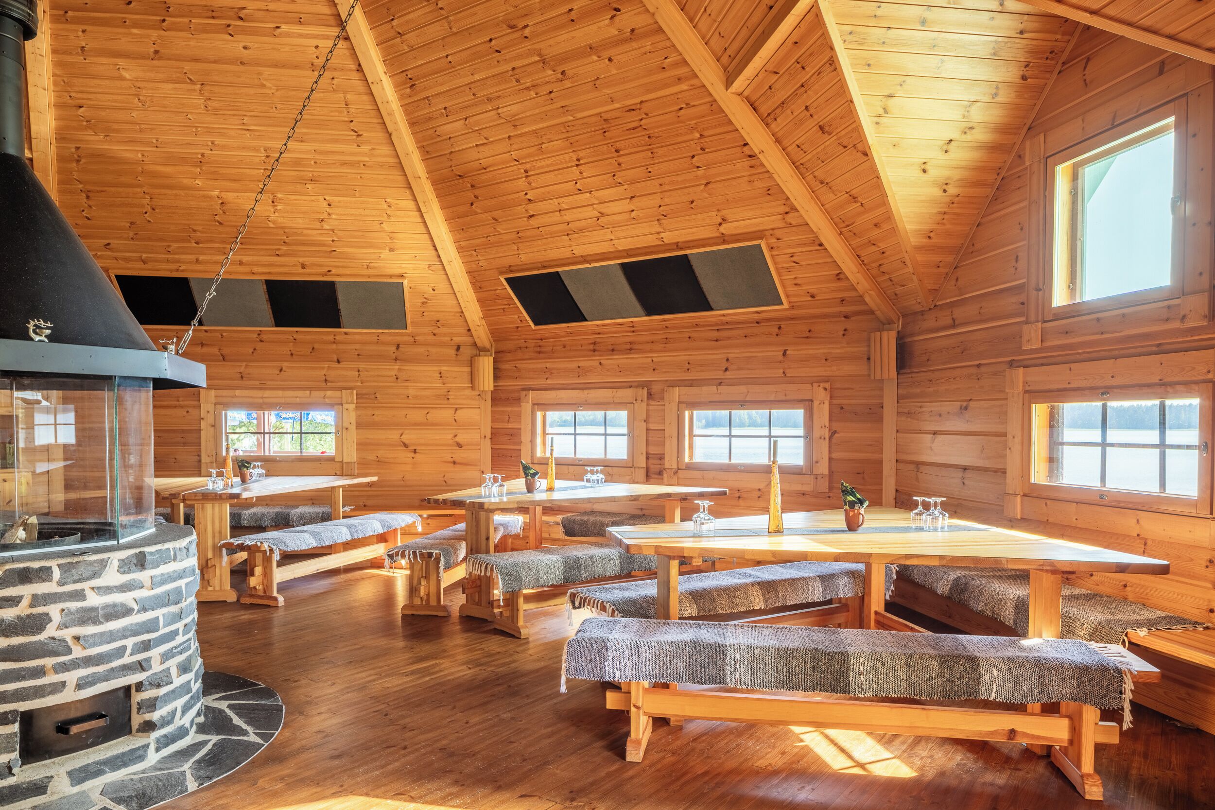 A log cabin interior with a stone fireplace, wooden benches, and large windows.