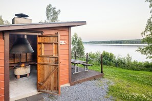 A rustic cabin with a large chimney, a grill, and a wooden deck overlooking a lake. - Hapimag Resort Punkaharju (Savonlinna)