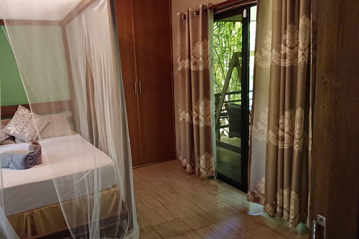 A bedroom with a bed under a mosquito net, a wooden wardrobe, and a view of a balcony with curtains.