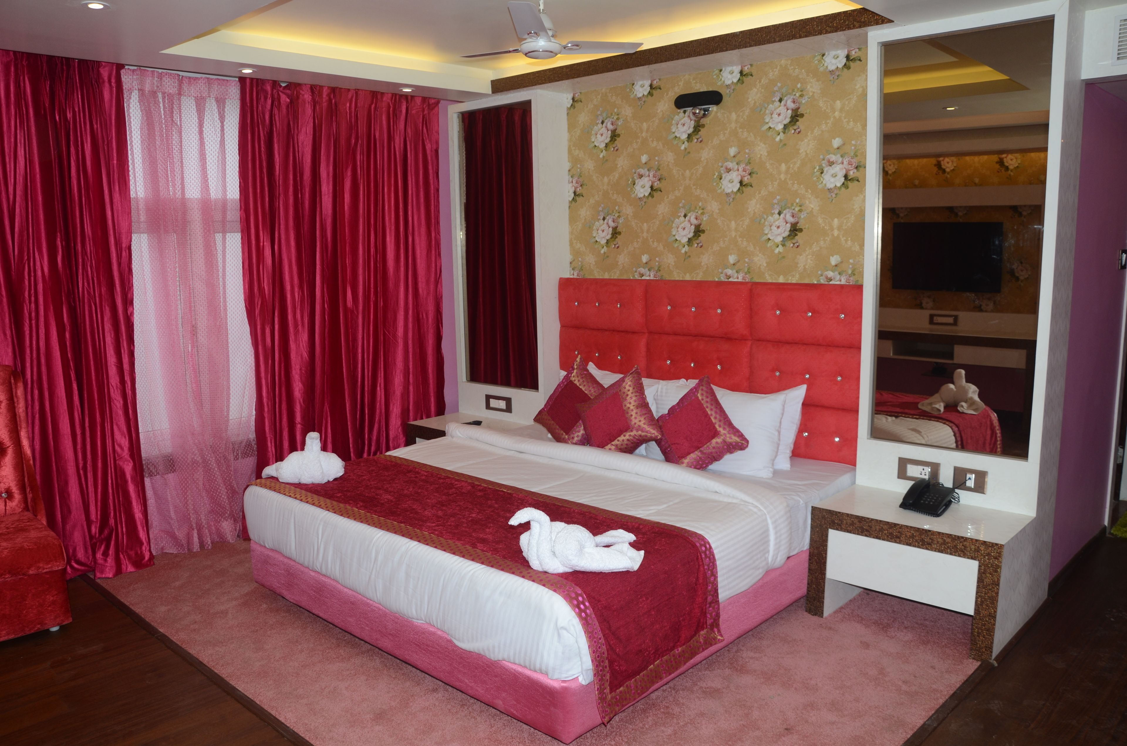 A hotel room with a large bed, red curtains, a red headboard, and a mirror.